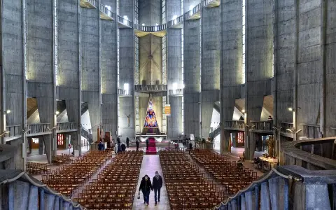 Intérieur de la cathédrale de Royan, avec ses murs en béton brut, ses rangées de chaises et son vitrail coloré au fond.