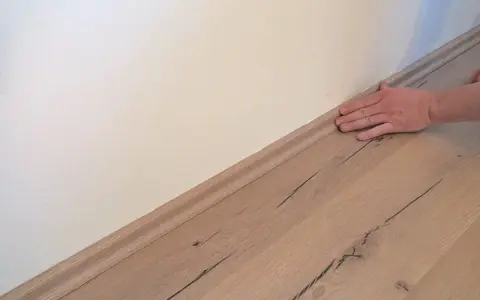 Hand drückt Sockelleisten an die Wand. Der Boden ist aus hellem Holz.