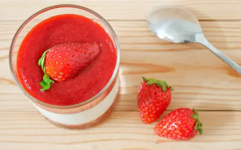 Un verre de panna cotta avec une fraise entière sur un coulis de fraise. Deux fraises fraîches et une cuillère à côté.