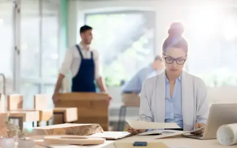 Kobieta w okularach analizuje dokumenty przy biurku, podczas gdy w tle mężczyzna w fartuchu pracuje przy drewnie. To jej staż z urzędu pracy.