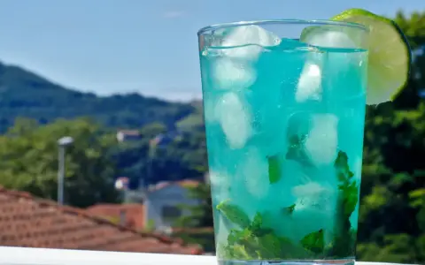 Orzeźwiający drink z blue curacao i sprite, z miętą i lodem, ozdobiony plasterkiem limonki, na tle zielonych wzgórz.