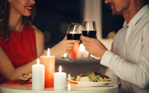Romantyczna kolacja przy świecach. Para wznosi toast kieliszkami czerwonego wina, uśmiechając się do siebie.