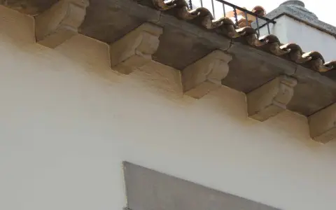 Detalle de qué es una cornisa, con ménsulas de piedra decorativas bajo tejas curvas y una barandilla metálica.