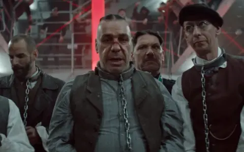 Rammstein - Deutschland: mężczyźni w łańcuchach, w tle czerwone światła i ludzie na schodach.