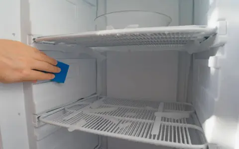 A hand uses a blue sponge to clean ice buildup inside a fridge freezer, showing a method for how to defrost fridge freezer without turning it off.