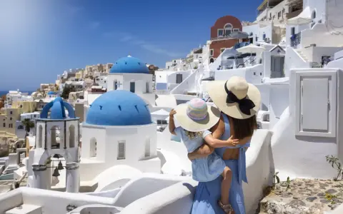Una madre abbraccia la sua bambina, ammirando il panorama di Santorini, Grecia. Le cupole blu brillano sotto il sole, mentre le case bianche si aggrappano alla collina.