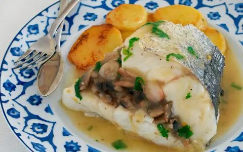Deliciosa merluza con gambas y champiñones, servida con patatas doradas en un plato azul y blanco.