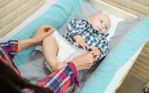 Mama sprawdza, kiedy zmienić rozmiar pieluchy maluszkowi. Dziecko leży na przewijaku w kratkę.