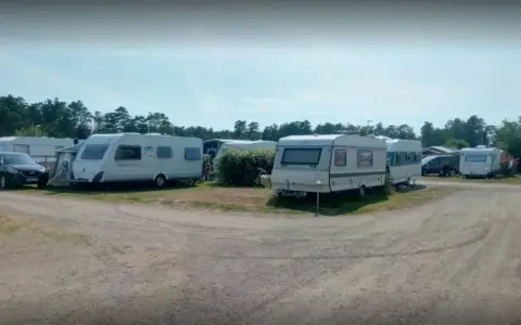 Ställplatser med husvagnar och bilar på Olles camping. Träd och blå himmel syns i bakgrunden.
