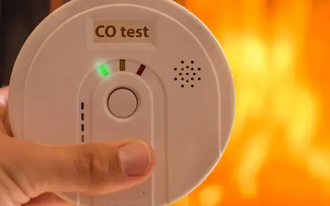 Detektor czadu z zieloną diodą i napisem "CO test" na tle płomieni.