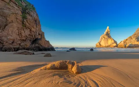 Goldene Sanddünen und Felsen am Praia da Ursa im warmen Abendlicht.