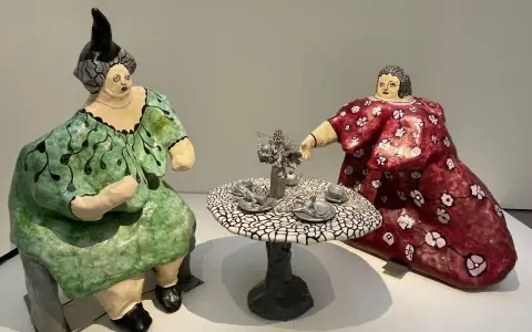 Dos figuras voluminosas, al estilo de Niki de Saint Phalle, sentadas a una mesa de té. Una viste de verde, la otra de rojo.
