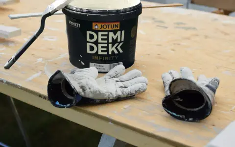 Förbereder för att måla utomhus på våren. En burk Jotun Demi Dekk, pensel och slitna handskar ligger på ett träbord.