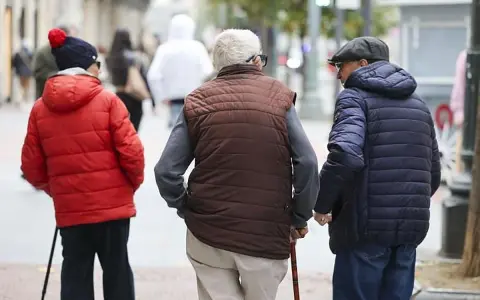Tres personas mayores caminan por la calle, una con bastón, reflexionando sobre la **pensión mínima**.