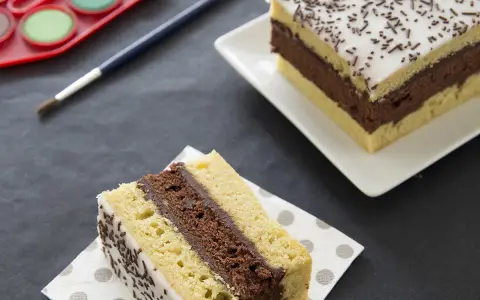 Une tranche de recette napolitain maison, avec glaçage blanc et vermicelles au chocolat, sur une serviette à pois. Un autre morceau est visible en arrière-plan.