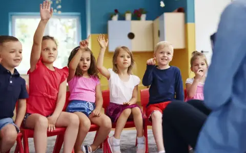 Dzieci na czerwonych krzesłach podnoszą ręce, jakby odpowiadały na pytanie, czy zerówka jest obowiązkowa.