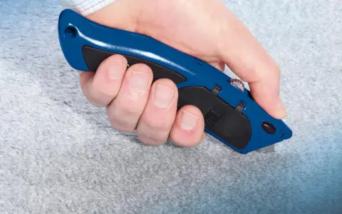 Hand hält blauen Teppichmesser, um grauen Teppich zu schneiden. Präzises Arbeiten beim Teppich verlegen.