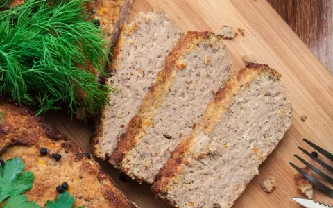 Pokrojony pasztet drobiowy z wątróbką, ozdobiony koperkiem i natką pietruszki, czeka na desce do krojenia.