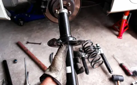 Mano guantata tiene un ammortizzatore nuovo, pronto per sostituire quelli scarichi dell'auto. Attrezzi e molle a vista.