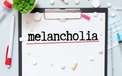Na kartce widnieje napis "melancholia". Wokół leżą tabletki, strzykawka i maseczka, sugerując walkę z tym stanem.