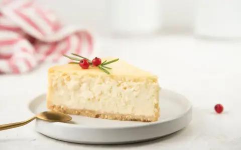 Una fetta di cheesecake con yogurt greco, guarnita con rosmarino e bacche rosse, su un piatto bianco con cucchiaino dorato.