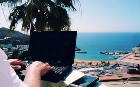 Praca na Cyprze: człowiek pisze na laptopie z widokiem na morze i palmę. Idealne miejsce na zdalną pracę.