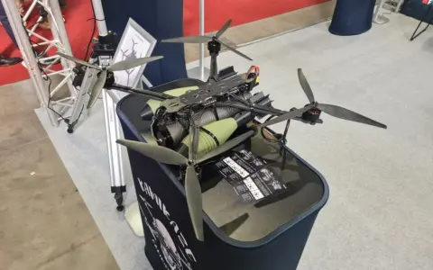 Ich sehe einen großen, schwarzen Quadrocopter mit grünen Zylindern, der auf einem Tisch steht. Die Propeller sind aus Carbon.