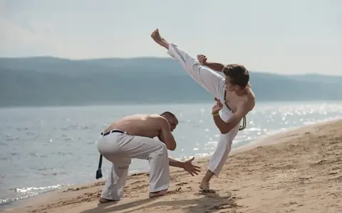 Dwóch mężczyzn ćwiczy capoeirę na plaży. Jeden wykonuje wysokie kopnięcie, drugi jest w pozycji obronnej. Widać ich płynny ginga styl gry.