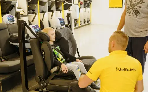 Chłopiec siedzi w foteliku samochodowym, a mężczyzna w żółtej koszulce z napisem "fstelik.info" wyjaśnia mu, od kiedy dziecko bez fotelika jest bezpieczne.