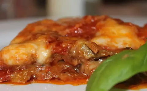 Deliciosa porción de parmigiana di melanzane, con capas de berenjena, salsa de tomate y queso fundido, adornada con una hoja de albahaca fresca.