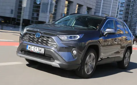 Ciemnoszary Toyota RAV4 Hybrid pędzi po mieście, odbijając w lustrach budynki i niebo.