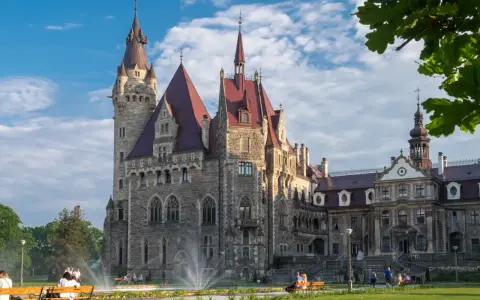 Imponujący zamek a pałac z kamienia, z wieżami i czerwonymi dachami, otoczony zielonym parkiem, gdzie odpoczywają ludzie.