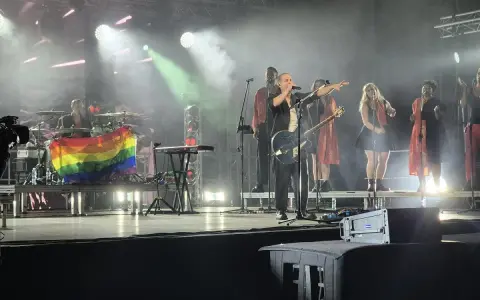 Wokalista z gitarą na scenie, obok chórki i perkusista z tęczową flagą. Koncert podczas festiwalu trzech kultur.
