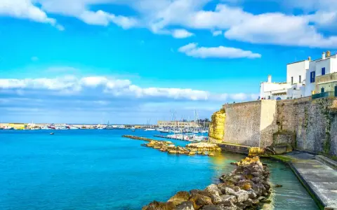 Un incantevole cammino del Salento: un porto con barche a vela, case bianche su scogliere dorate e un mare azzurro sotto un cielo limpido.