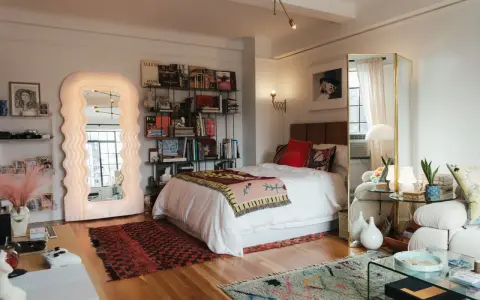 Ideas para decorar habitación estudio: cama blanca, espejo ondulado, estanterías con libros y arte, alfombras coloridas y un sofá acogedor.