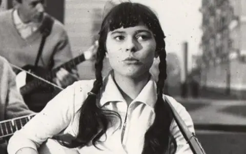 Młoda Karin Stanek z warkoczami śpiewa, grając na gitarze. W tle zespół muzyczny.