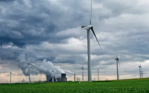 Windräder auf grüner Wiese unter dramatischem Himmel, im Hintergrund rauchende Industrieanlagen. Ein Symbol für Investitionen in erneuerbare Energien in Deutschland.