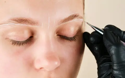 Une main gantée dessine des lignes blanches sur un sourcil pour un microblading poil à poil.