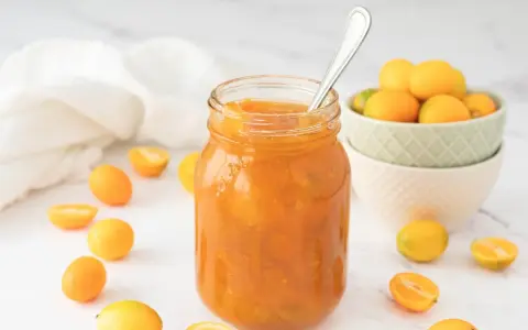Tarro de mermelada de kumquat casera, rodeado de kumquats frescos y cortados. Una cuchara plateada reposa en el tarro.