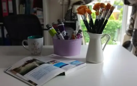 Pędzelki malarskie w dzbanku, tubki farb w fioletowym wiaderku, kubek z napisem i otwarta książka.