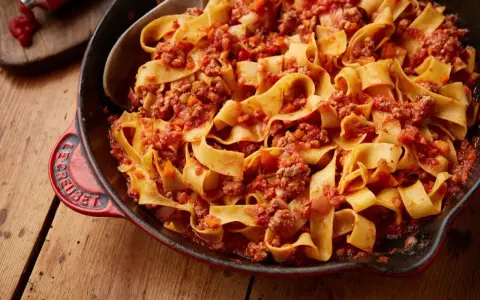 En stor portion pasta bolognese med breda bandnudlar och en mustig köttfärssås i en röd gjutjärnspanna.
