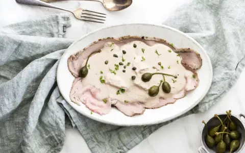 Eleganckie vitello tonnato z kremowym sosem, kaparami i natką pietruszki na białym talerzu, obok srebrnych sztućców i lnianej serwetki.