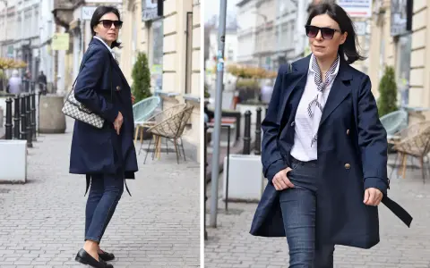 Elegancka kobieta w granatowym trenczu damskim, białej bluzce w paski i jeansach spaceruje po brukowanej ulicy.