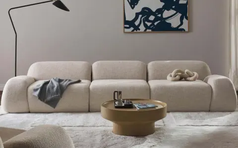Divano modulare in tessuto bouclé color crema, con cuscino decorativo a forma di catena. Il materiale del divano è morbido e invitante, perfetto per un salotto moderno.