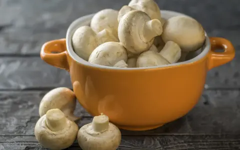 Pomarańczowa miseczka pełna świeżych pieczarek, jakby w ciąży z nowymi smakami. Kilka pieczarek leży obok na drewnianym blacie.