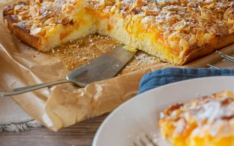 Duże ciasto z brzoskwiniami i migdałami, posypane cukrem pudrem, na papierze do pieczenia.