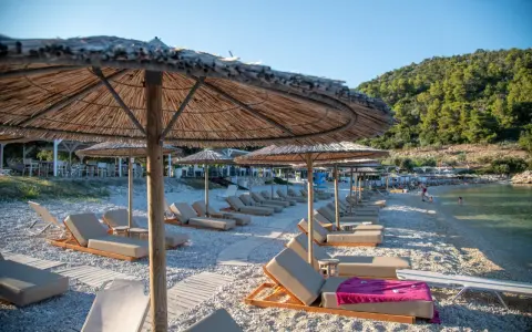 Alonissos spiagge: lettini e ombrelloni di paglia su una spiaggia di ciottoli, con il mare cristallino e la collina verde sullo sfondo.