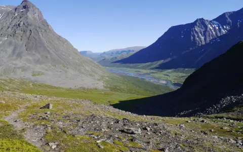 Vacker vy över Sareks nationalpark med Rapadalen i fjärran. En flod slingrar sig genom en grön dal omgiven av höga, steniga berg under en klarblå himmel.