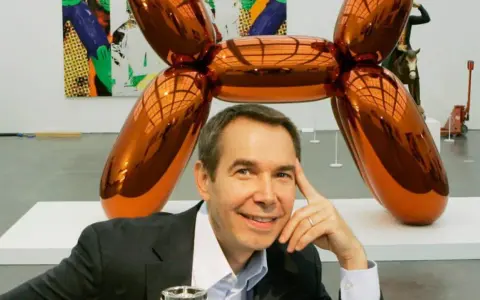 Jeff Koons posa junto a su icónica escultura perro globo naranja brillante en una galería de arte.