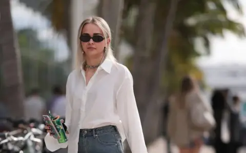 Donna con occhiali da sole, collana, camicia bianca e jeans, tiene in mano una lattina verde e un telefono.